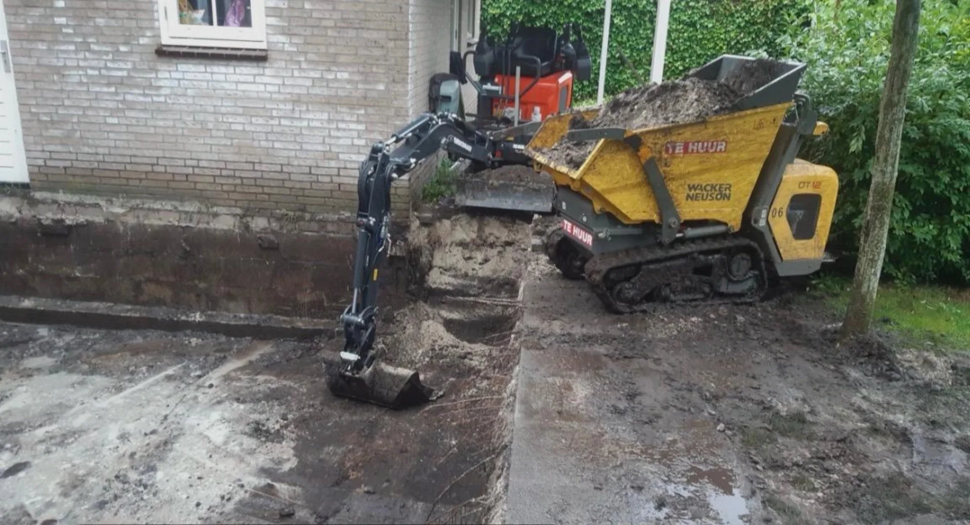 Minigraver graaft een sleuf langs de gevel en laadt grond in een rupsdumper